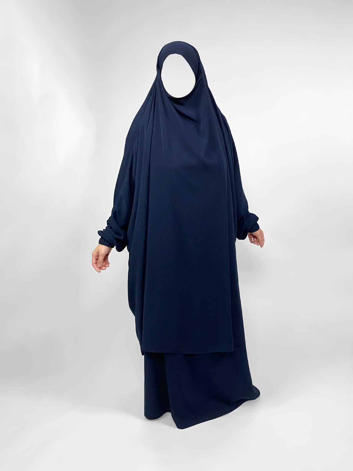 A navy two-piece jilbab against a pale studio backdrop: a three-quarter over-piece covering the head and falling to roughly the knee, worn over a long skirt that reaches the floor.